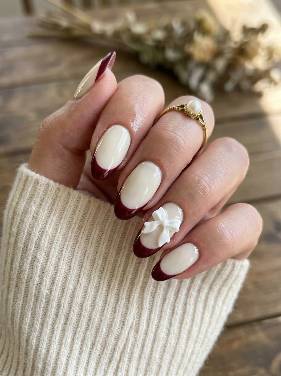 Almond nails with milky white base, deep burgundy French tips, and a 3D white bow charm on the ring finger.