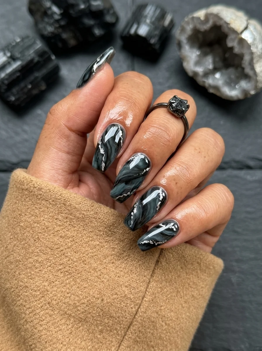 Long almond nails with a black base and silver-white marble swirl with glitter accents.