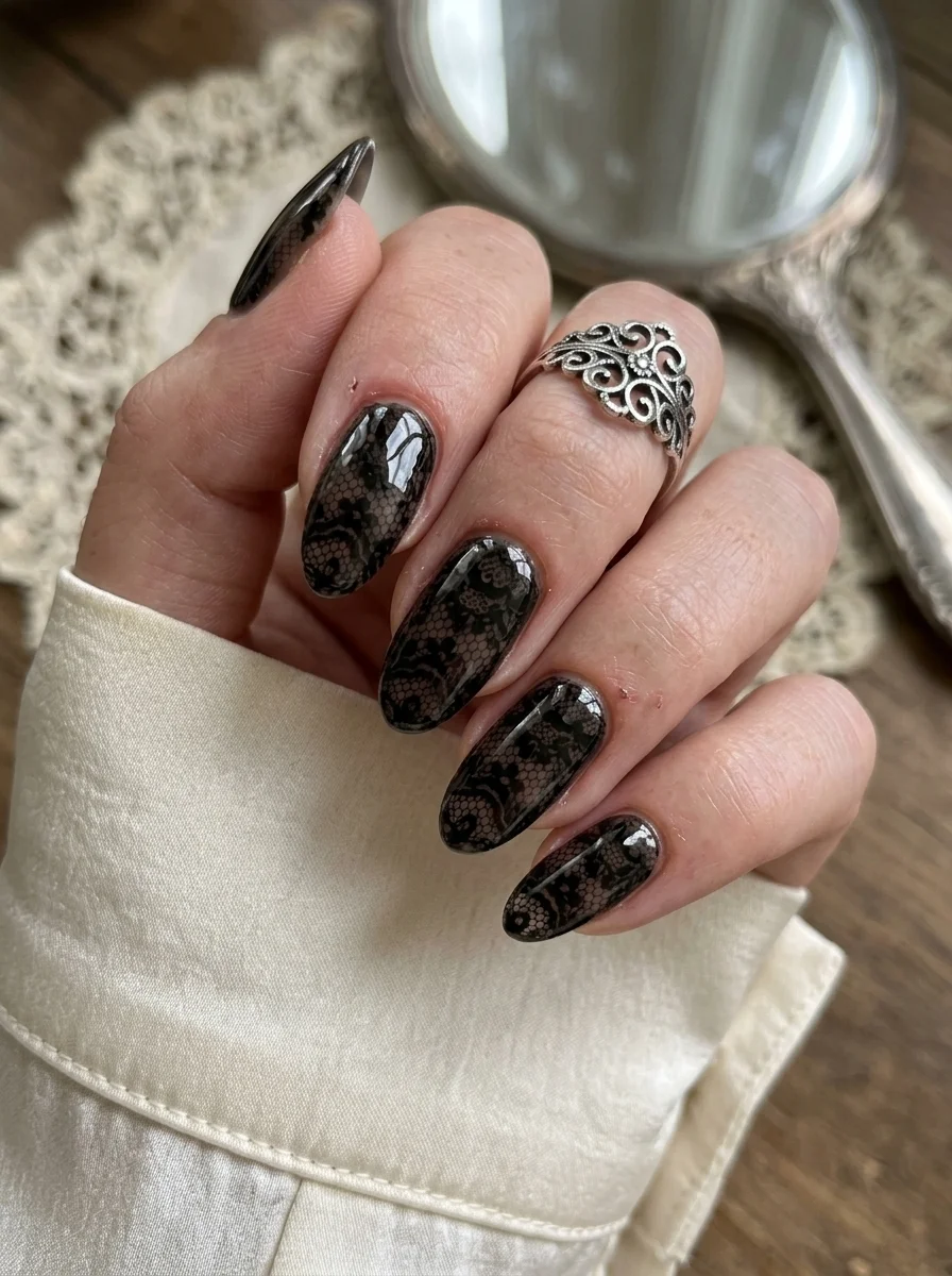 Long almond nails with a sheer nude base and intricate black lace patterns painted across them.